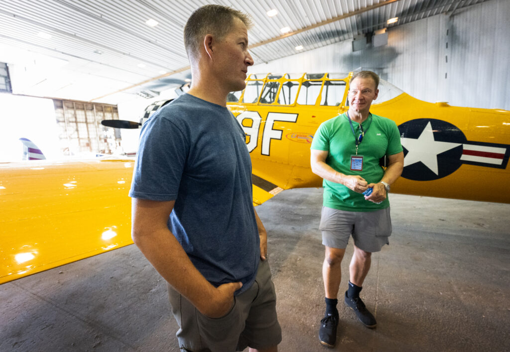 Two pilots in T-shirts stand near a yellow plane in a hangar. 