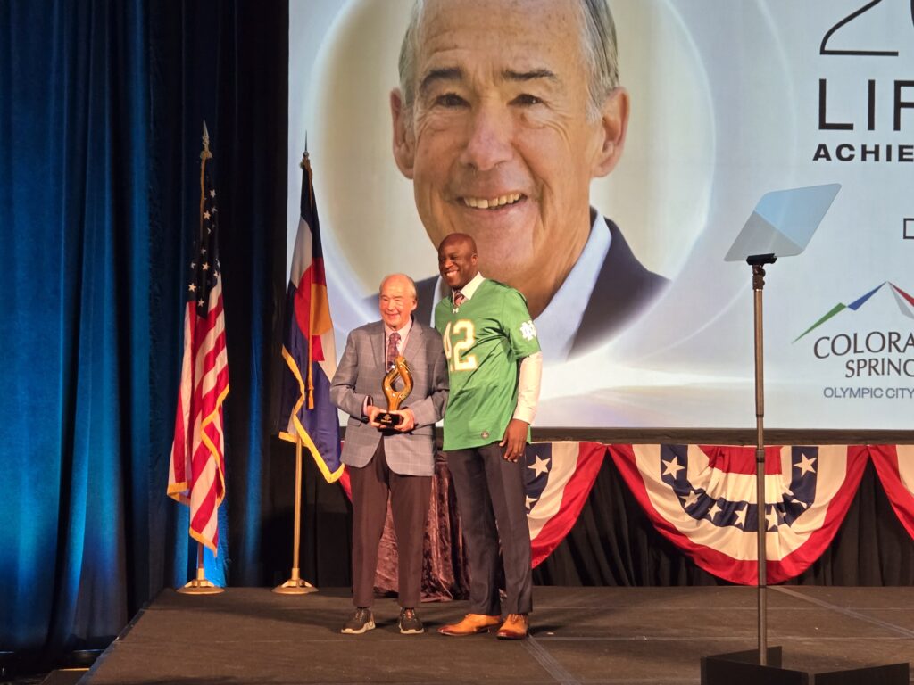 David Lord and Yemi Mobolade stand on stage together. Lord is holding the Spirit of Colorado Springs Lifetime Achievement Award.