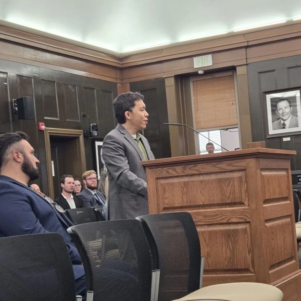 Kaname Kuniyuki standing behind a podium speaking to off-screen members of the Colorado Springs City Council.