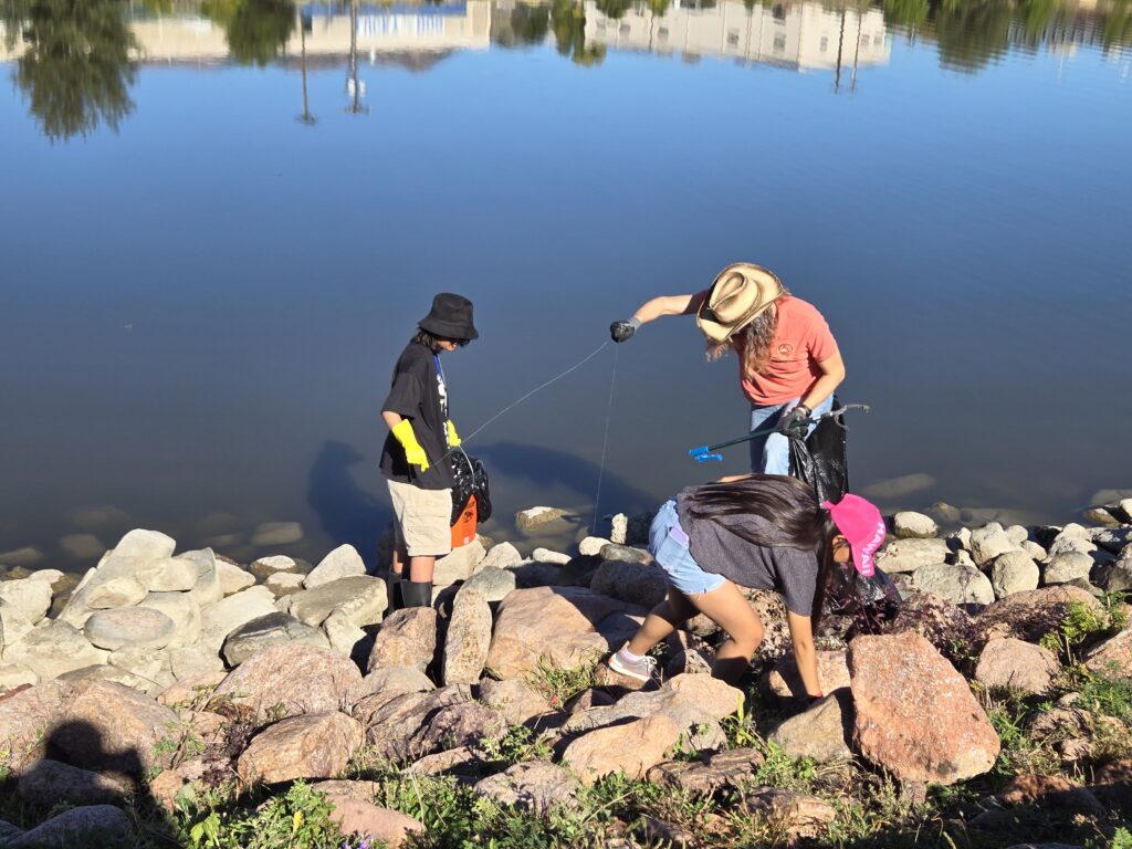 Two high schoolers pick up fishing line from rocks on the shore of a reservoir, while a third student does other cleanup.