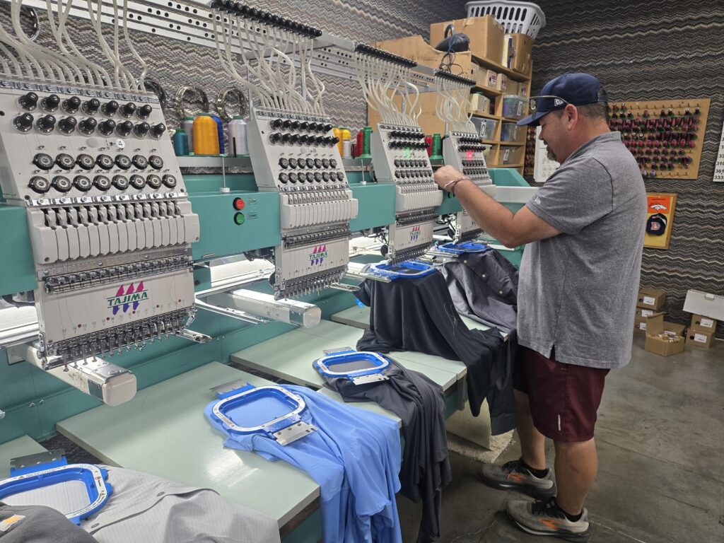 Shirt Stop employee Eric Davenport oversees the custom embroidering of an order on Friday, Oct. 3, 2025. He stands at a row of several automated sewing machines as they embroider shirts.
