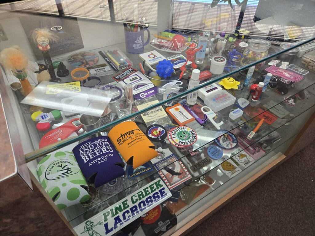 A glass counter is filled with various types of promotional items that can be customized by Shirt Stop. On display are items including koozies, bumper stickers, patches, lip balms, bottle openers and more.