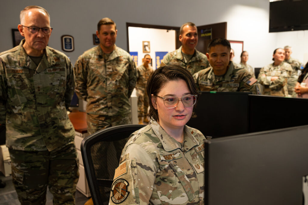 An Air Force reservist in fatigues sits at a computer console while running satellites. Officers stand behind her. 